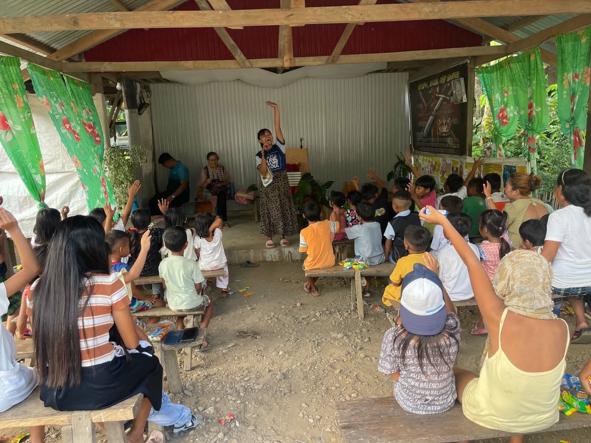 Children seated on small chairs facing a speaker in a covered gathering space