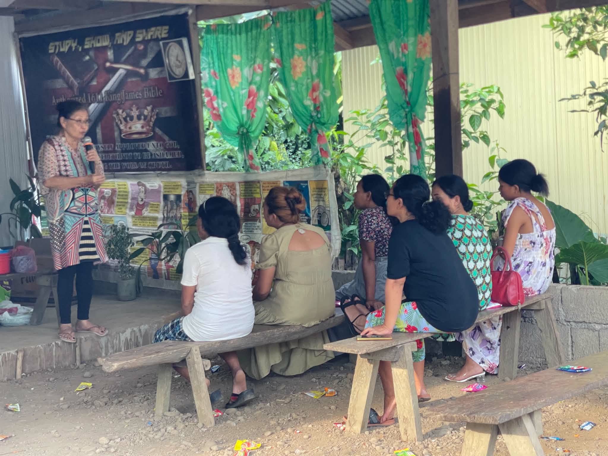 An adult speaking to a seated group in a shaded gathering space