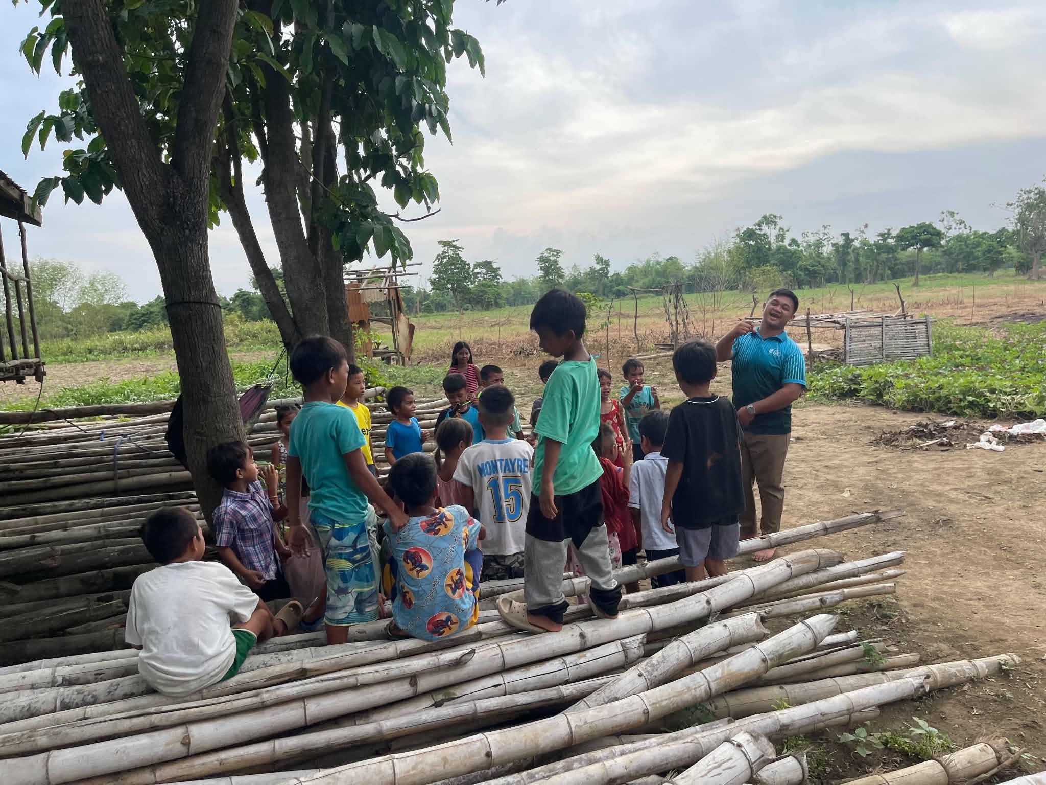 Children seated outdoors near a tree while adults stand nearby
