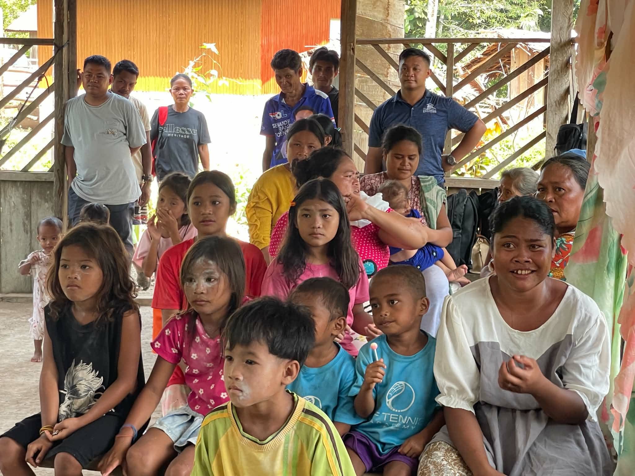 Children gathered with adults inside an open wooden shelter